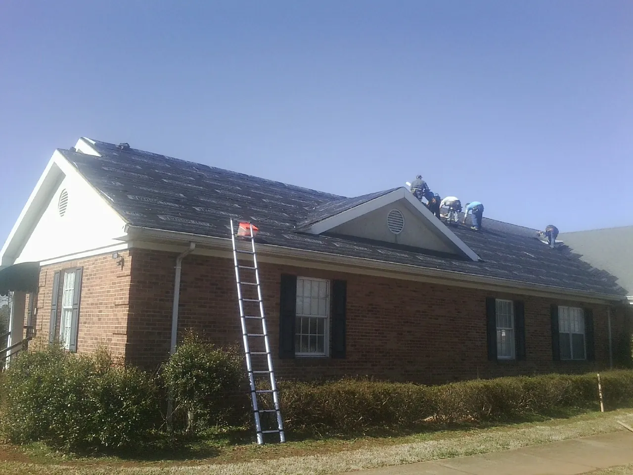 Crew tearing shingles off a brick ranch roof with a ladder set — Metal Roof Repair work by our crew in Pepperell