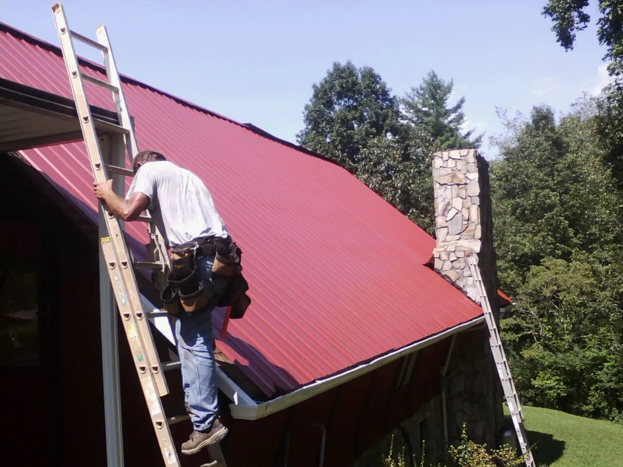 Roofer on a ladder beside a red metal roof and stone chimney — Soffit & Fascia Repair by Roofing Brothers in Pepperell